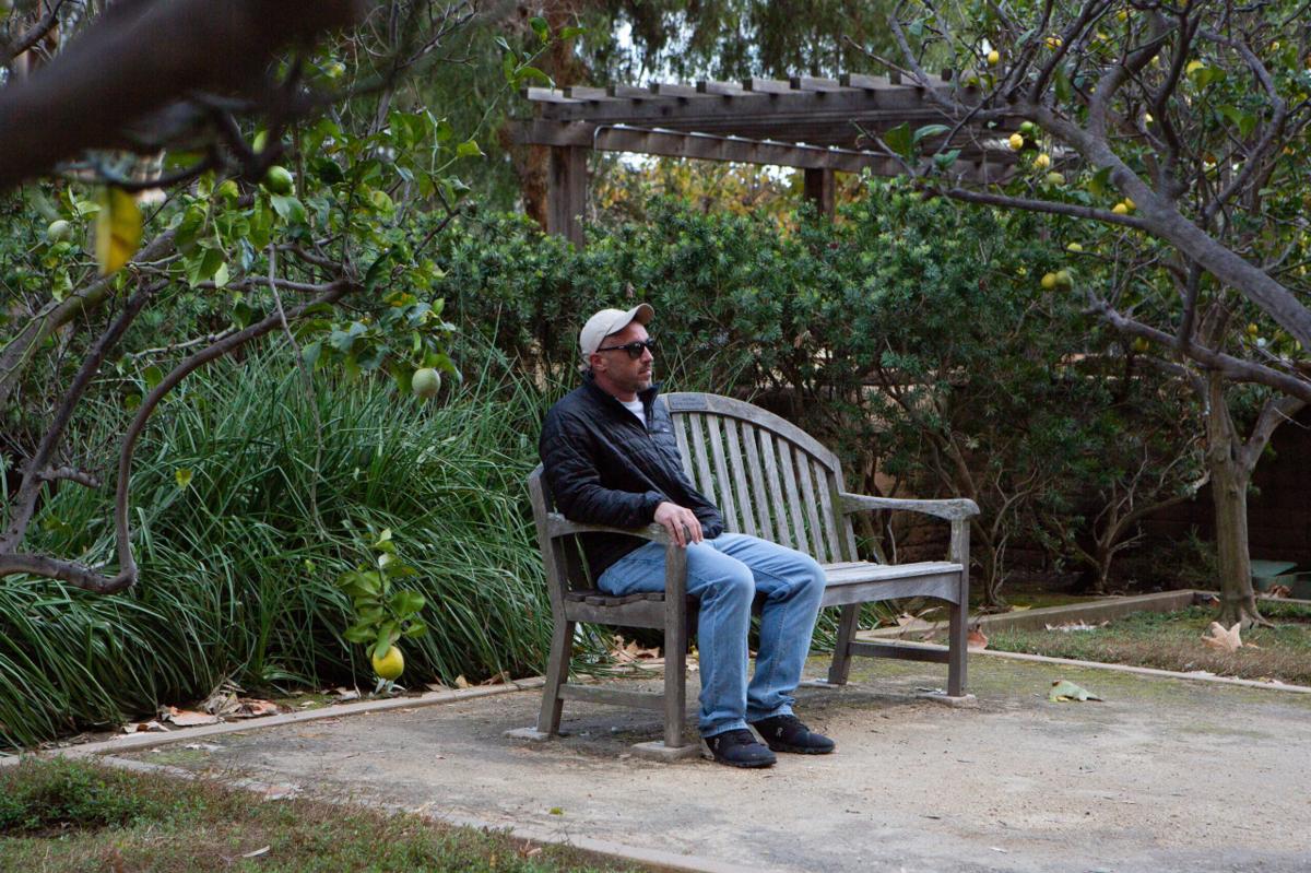 Rob Reynolds, wearing a baseball hat and sunglasses, sits on a wooden bench in the West LA VA rose garden.