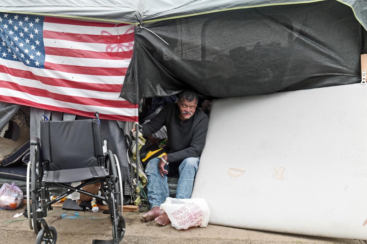 An unhoused man sits in a makeshift tent on a sidewalk, flanked by an American flag, a mattress, and his wheelchair.